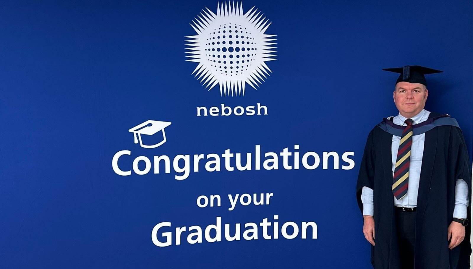 A photo of ACT graduate Martyn Hill wearing a graduation cap and gown, next to a large backdrop that reads 'NEBOSH -Congratulations on your Graduation' 